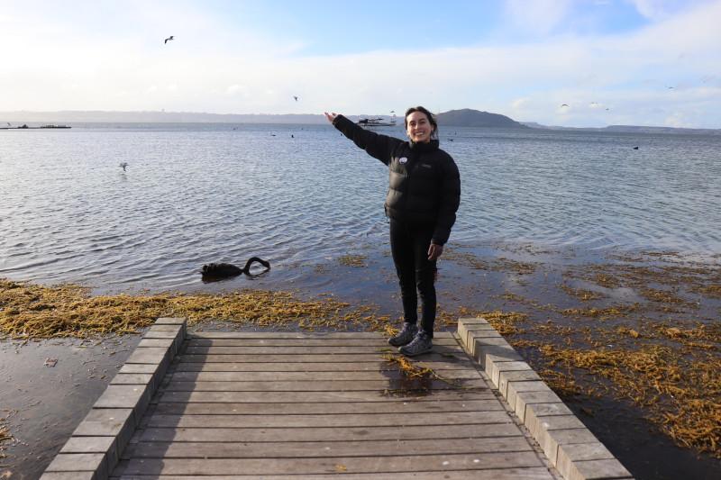 International student Daniela stands on the edge of a wharf and points out at the ocean and seabirds